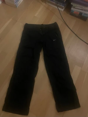 Svarta byxor från Nike - Säljer ett par svarta byxor från Nike med raka ben och dragkedja samt dubbla knappar i midjan. Byxorna har fickor fram och en liten vit Nike-logga på ena sidan. Materialet ser ut att vara bomull eller en bomullsblandning.