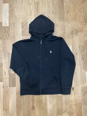Svart hoodie från Polo Ralph Lauren - (Priset går att diskuteras) Klassisk svart hoodie från Polo Ralph Lauren med dragkedja, huva med dragsko och kängurufickor. Liten vit logga broderad på bröstet. Tillverkad i mjukt bomullsmaterial som känns skönt mot huden. Perfekt för en avslappnad och stilren look.