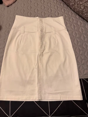 Vit pennkjol med hög midja - Säljer en stilren vit pennkjol med hög midja och bälteshällor. Kjolen har en rak passform och är sydd i ett mjukt bomullsliknande material. Perfekt att matcha med en crop top eller en oversized tröja för en trendig look.