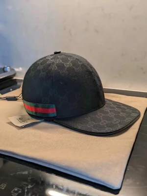 Svart Gucci keps med GG-mönster - Snygg svart keps från Gucci med klassiskt GG-monogram över hela kepsen. På sidan finns en grön och röd rand som ger en ikonisk touch. Kepsen har böjd skärm och justerbar rem bak. Materialet är canvas med exklusiv känsla.