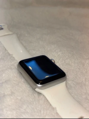Apple Watch med vitt sportarmband - Snygg Apple Watch med rektangulär svart pekskärm och aluminiumboett i silver. Klockan har ett vitt sportarmband i silikon som är bekvämt att bära hela dagen. Perfekt för dig som vill ha koll på notiser, träning och hälsa direkt på handleden. Kommer också med ett svart band 