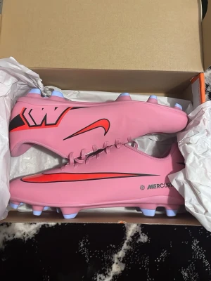 Nike Mercurial rosa fotbollsskor - Använt bara 1 gång. Snygga rosa Nike Mercurial fotbollsskor med röda och svarta detaljer samt vita dobbar. Skorna har en slimmad form och snörning för bra passform. Tillverkade i syntetmaterial som ger bra bollkontroll och grepp på planen. Perfekta för dig som vill sticka ut på fotbollsplanen.