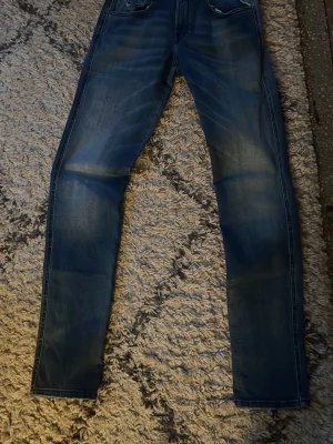 Blå slim jeans från Replay - Säljer ett par helt nya blå slim fit jeans från Replay med snygga slitningar och klassiska fem fickor. Jeansen har coola detaljer på bakfickorna och är tillverkade i denim. Perfekta för dig som gillar en modern och avslappnad stil.