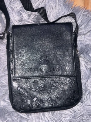 Svart axelväska Philipp Plein - Säljer en svart axelväska från Philipp Plein i läderimitation med präglade dödskallar och logotyp på framsidan. Väskan har justerbar axelrem och praktisk ficka med lock. Perfekt för dig som gillar edgy detaljer och streetstyle.