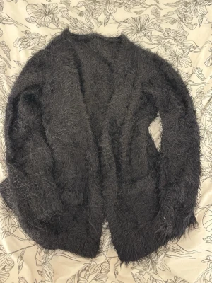 Svart fluffig stickad kofta - Säljer en supermjuk och fluffig svart stickad kofta utan knappar. Koftan har lång ärm och öppen front, perfekt för lager på lager och en chill look. Materialet är lurvigt och ger en cozy vibe.