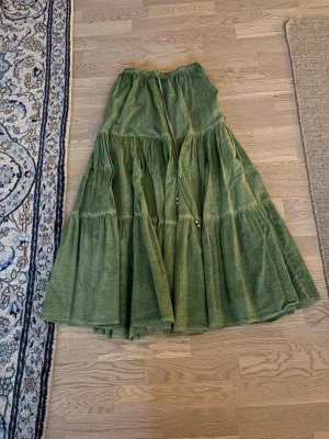 Grön långkjol i linne med snörning - Säljer en flowy grön långkjol i linne med snygg snörning i midjan och pärldetaljer på snörena. Kjolen har flera volanger och en avslappnad passform som ger en bohemisk vibe. Perfekt för dig som gillar naturfärger och sköna material.