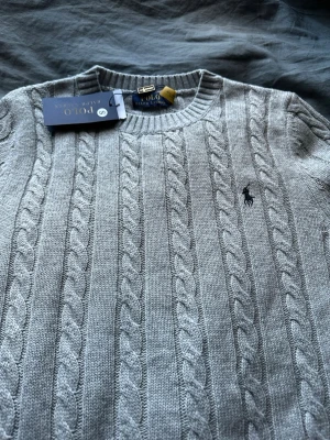 Grå kabelstickad tröja Polo Ralph Lauren - Snygg grå kabelstickad tröja från Polo Ralph Lauren med rund halsringning och klassisk broderad logga på bröstet. Tröjan har ribbade muddar och är tillverkad i mjukt material som passar perfekt till lager-på-lager-stil. STORLEK SMALL (S) alla frågor är välkomna 