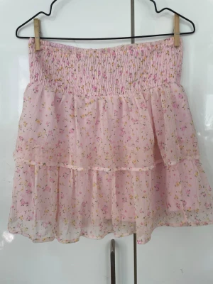 Rosa blommig volangkjol Vero Moda - Supersöt rosa kortkjol från Vero Moda med små blommor i gult och rosa. Kjolen har volanger och smock i midjan för en bekväm passform. Perfekt för vår och sommar, lätt och luftig känsla.