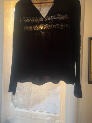 Svart blus med spetsdetaljer - Snygg svart blus med långa ärmar och v-ringning. Blusen har fina spetsdetaljer över bröstet och knytband vid halsen. Tillverkad i ett tunt, lätt material som ger ett luftigt intryck. Perfekt att styla med jeans eller kjol.