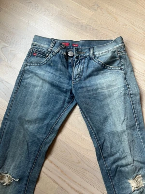 Lågmidjade bootcut jeans med hål  - Säljer nu mina favorit jeans med slitningar och hål på båda knäna. Jeansen har klassisk femficksmodell,  bootcut och detaljer i rött vid fickorna. Helt perfekta! Säljer pga de har blivit försmå.