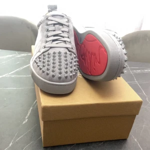 Gråa Louboutin sneakers med nitar - Säljer ett par gråa sneakers från Christian Louboutin med coola nitar på tån och sidorna. Skorna har den ikoniska röda sulan och är gjorda i mocka. Klassisk snörning och låg profil ger en clean look.