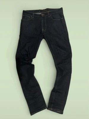 Mörkblå jeans från Nudie Jeans - Säljer ett par mörkblå jeans från Nudie Jeans med klassisk femficksmodell och kontrastsömmar. Jeansen har rak passform och är tillverkade i kraftig denim med Nudie Jeans signaturbrodyr på bakfickan. Perfekta för en stilren och tidlös look.