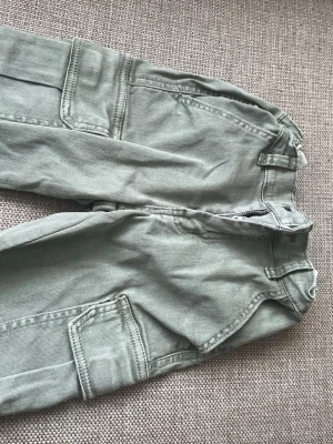 Gröna cargobyxor från D*M*D - Snygga cargobyxor i ljusgrönt från D*M*D med klassiska stora fickor på sidorna och dragkedja fram. Tillverkade i ett slitstarkt bomullstyg som ger en avslappnad och trendig look. Perfekta för dig som gillar streetwear och praktiska detaljer. Obs finns fläck som ni ser märks dock inte så mkt , vet inte om den går bort i tvätten .