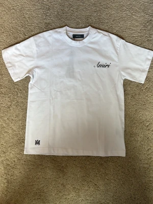 Vit Amiri t-shirt med logga - Snygg vit t-shirt från Amiri med svart logga på bröstet och en liten detalj nere vid fållen. Klassisk passform och rund halsringning. Tillverkad i mjuk bomull som känns skön mot huden. Perfekt för dig som gillar stilrena plagg med exklusiv känsla. Den är lite större i storleken och lite mer baggy. 