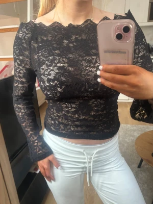 Svart spetsblus med lång ärm - Snygg svart blus i spets med lång ärm och transparent design. Blusen har en vacker vågig kant vid halsen och är figurnära i modellen. Perfekt för dig som vill ha en trendig och elegant topp med det lilla extra.