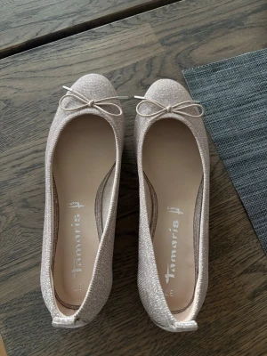 Glittriga ballerinaskor från Tamaris - Säljer ett par glittriga ballerinaskor från Tamaris i beige med en söt rosett framtill. Har aldrig använt dom❤️Helt nya! glittriga ballerina lite champagne liknande färg. Dom är ganska stora i storleken jag brukar ha 38/39 men dessa sitter perfekt 