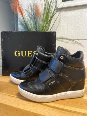 Svarta höga sneakers från Guess - Snygga svarta sneakers från Guess med dold kilklack. Skorna har vita sulor och Guess logo detaljer. Skorna har breda kardborreband och logga på sidan. 