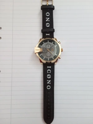 Svart klocka/armbandsur - Snygg svart klocka med bred svart rem i syntetmaterial och vita bokstäver. Gulddetaljer på sidan och texten 'HOMME' på skyddet. Köptes på NewYorker. Använd ett mycket fåtal gånger, och har därför inga spår av användning. Batteriet fungerar inte.