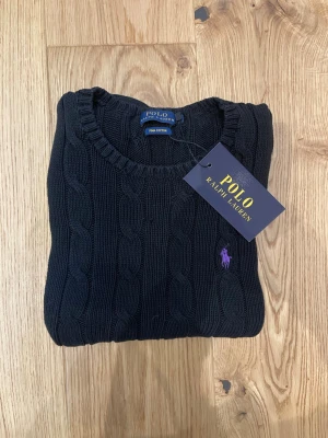 Mörkgrå kabelstickad tröja Polo Ralph Lauren storlek S - Kabelstickad mörkgrå tröja från Polo Ralph Lauren med lila broderad logga på bröstet. Tröjan har rund halsringning, långa ärmar och ribbade muddar. Jag säljer då den inte passar mig. Skriv vid minsta funderingar. MVH Tim.