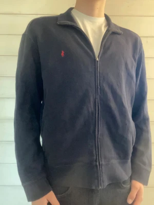 Mörkblå ziptröja från Ralph Lauren - Mörkblå ziptröja från Ralph Lauren med röd broderad logga på bröstet. Tröjan är i fint skick förutom nere på tröjan lite vid dragkedjan där är den lite sliten. Skriv gärna vid några frågor!