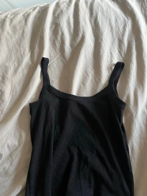Svart ribbad linnetopp från Vero Moda - Svart ribbad linnetopp från Vero Moda med smala axelband och enkel, stilren design. Perfekt att matcha med jeans eller kjol för en snygg look. Materialet är mjukt och stretchigt, vilket gör den bekväm att bära hela dagen.