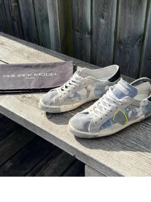 Philippe Model camo sneakers - Snygga sneakers från Philippe Model med grått och blått camomönster, vita detaljer och en gul markerad logga på sidan. Skorna har vita snören, svart häl och är tillverkade i mocka och textil. Perfekta för dig som gillar streetwear och unik design.