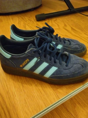Adidas Spezial blå mocka sneakers - Säljer ett par Adidas Spezial sneakers i blå mocka med ljusblå detaljer och klassiska tre ränder på sidan. Skorna har mörkblå snören, gummisula och guldfärgad Spezial-logga på sidan. Perfekta för dig som gillar retrostil och streetwear. Endast använda enstaka gånger, säljer pga att jag ej använt dem mycket. Nypris 1349kr