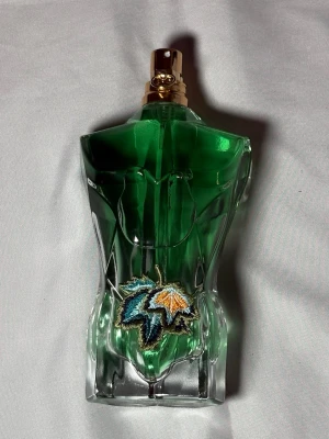 Grön parfym Jean Paul Gaultier Le Male - Unik parfymflaska från Jean Paul Gaultier Le Male, formad som en manlig torso i grönt glas med guldfärgad spraytopp. Flaskan har en broderad detalj med färgglada löv framtill, vilket ger en cool och kreativ look. Perfekt för dig som gillar statement-parfymer.