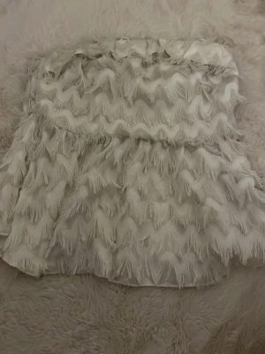 Vit tubtopp - Supersnygg vit tubtopp med fransdetaljer i vågmönster. Toppen är axelbandslös och har en elastisk smock baktill för skön passform. Perfekt för dig som vill sticka ut med en unik och trendig look.