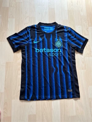 Inter Milan matchtröja Nike svart/blå - Säljer en Inter Milan matchtröja från Nike i svart och blått med vertikala ränder. Tröjan har korta ärmar, rund hals och tryckta loggor från Nike, Inter och Betsson Sport på bröstet. Perfekt för fotbollsträning eller att bära till vardags.