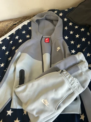 Nike grå hoodie och mjukisbyxor - Nike tech med hoodie och mjukisbyxor i ljusgrå och mellangrå toner. Hoodien har dragkedja, fickor och Nike-logga på bröstet. Byxorna har resår i midjan, sidofickor och vit Nike-logga. Perfekt för chill dagar hemma eller när du vill vara bekväm.
