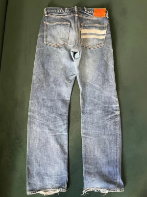 SKIT FETA MOMONTARO - Säljer ett par blå raka jeans klassisk femficksdesign och slitna detaljer vid bensluten. Jeansen har ljus tvätt, kontrastsömmar och tre beige ränder på ena bakfickan samt en orange patch i midjan. Perfekta för en avslappnad streetwear-look. Skriv gärna till mig innnan ni köper!!