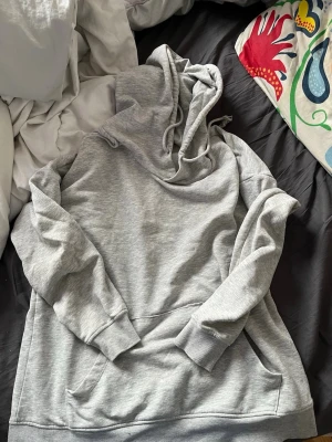 Grå hoodie med magficka - En klassisk grå hoodie med huva och dragsko samt stor magficka framtill. Tillverkad i mjukt sweatshirtmaterial, perfekt för chill dagar. Ribbade muddar vid ärmslut och nederkant för extra komfort. Det finns 4 SMÅ blå fläckar på tröjan som knappt syns.