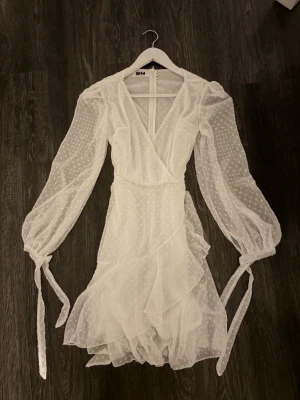 Vit Dennis Maglic klänning - Söt vit kortklänning med omlottmodell, volangdetaljer och långa transparenta ärmar. Klänningen har små prickar i tyget, knytband vid ärmsluten och v-ringad hals. Perfekt för sommarens alla tillfällen!