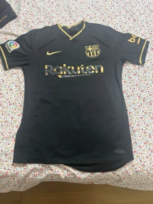 Svart FC Barcelona matchtröja Nike - Snygg svart FC Barcelona matchtröja från Nike med guldfärgade detaljer och loggor. Rakuten och Unicef tryck på fram- och baksidan, samt klubbmärke och Nike swoosh på bröstet. Korta ärmar och sportig passform, perfekt för fotboll eller träning.