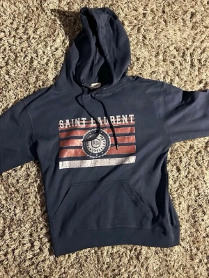 Saint Laurent hoodie - Mörkblå hoodie från Saint Laurent med tryck framtill i vitt och rött samt logga. Klassisk känguruficka och justerbar huva med snören. Mjuk bomullskänsla och avslappnad passform, perfekt för en cool och casual look.