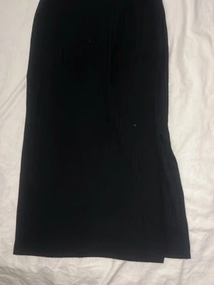Svart långkjol med slits - En stilren svart långkjol med slits på sidan. Kjolen har en rak passform och är tillverkad i ett mjukt, lätt material som faller snyggt. Perfekt att matcha med en crop top eller stickad tröja för en trendig look.