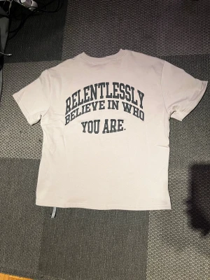 Beige t-shirt med tryck - Beige t-shirt från Zara med svart texttryck på ryggen: 'RELENTLESSLY BELIEVE IN WHO YOU ARE.' Klassisk rund halsringning och korta ärmar. Tillverkad i mjuk bomull för en skön och avslappnad känsla.