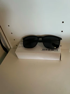 Svarta solglasögon med klassisk form - Snygga svarta solglasögon med klassisk, rak båge och mörka glas. Ramen är i plast och har en stilren look som passar till det mesta. Perfekta för soliga dagar och ger en cool vibe till din outfit.