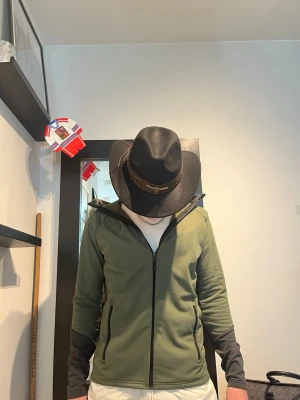 Grön vindjacka med huva från peak performance  - Snygg olivgrön vindjacka med svarta detaljer och dragkedja framtill. Jackan har huva och svarta ärmar samt fickor med dragkedja. Perfekt för dig som gillar en sportig och avslappnad stil. Modellen är 182 och väger 75kg