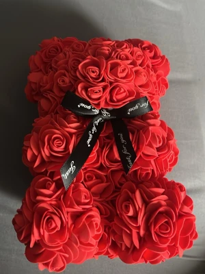 Rosbjörn med svart rosett - En dekorativ björnformad figur täckt av röda konstgjorda rosor och prydd med en svart rosett med texten 'Just for you'. Perfekt som present eller inredningsdetalj. Materialet är mjukt skum och plast. 