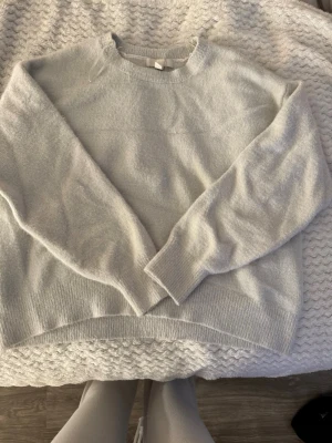 Ljusgrå/vit stickad tröja - Mysig ljusgrå/vit stickad tröja med rund halsringning och långa ärmar. Tröjan har ribbade muddar vid ärmslut och nederkant samt en något längre bakdel för en avslappnad look.