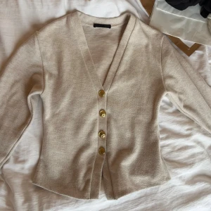 Beige kofta med guldknappar - Stilren beige kofta med fyra stora guldknappar framtill och djup V-ringning. Koftan har lång ärm och är gjord i ett mjukt, tunt stickat material som känns skönt mot huden. Perfekt att slänga över en topp för en enkel men snygg look.