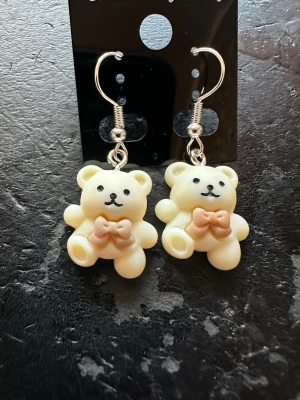 Söta björnhänge örhängen - Ett par örhängen med krämvita björnar i plast, dekorerade med ljusbruna rosetter. Hängena har krokar i silverfärg och björnarna har svartmålade ansikten. Perfekta för dig som gillar gulliga accessoarer med lekfull stil.