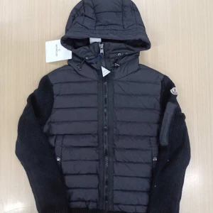 Svart Moncler cardigan jacka med huva - Svart cardigan jacka från Moncler med quiltad framsida och huva. Ärmarna är stickade och har en patchficka på ena armen med Moncler-logga. Jackan har dragkedja framtill och ribbade muddar. Perfekt för dig som vill ha en snygg och sportig look.