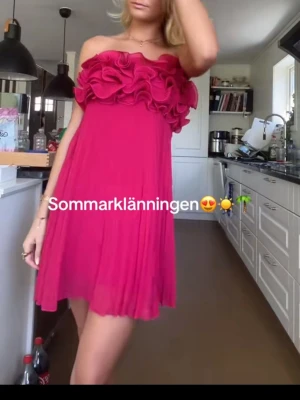 Axelbandslös rosa kortklänning - Snygg axelbandslös kortklänning i stark rosa färg med volangdetaljer upptill. Klänningen har ett plisserat tyg som ger ett luftigt intryck och passar perfekt för varma dagar. Materialet ser ut att vara lätt och skönt, troligen polyester.