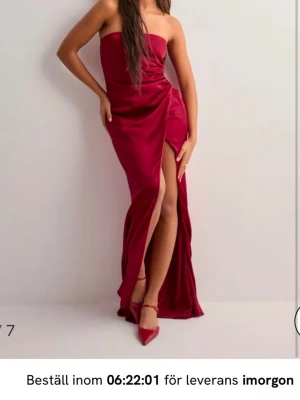 Axelbandslös vinröd maxiklänning - Snygg axelbandslös maxiklänning i vinrött med hög slits framtill. Klänningen har en figurnära passform och elegant drapering vid höften. Perfekt för dig som vill sticka ut på festen.