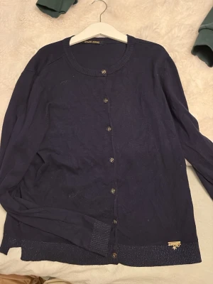 Mörkblå kofta från Gaudi Jeans - Mörkblå tunnstickad kofta från Gaudi Jeans med rund halsringning och knappar framtill. Långa ärmar och ribbade muddar vid ärmslut och nederkant. Liten metallogo vid nederkanten ger extra detalj. Den säljs inte mer och är lite skrynklig men den har inte används så mycket 