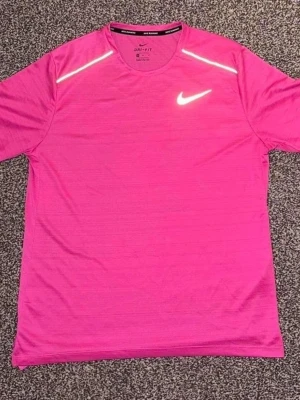 Rosa Nike Dri-Fit t-shirt - Snygg rosa Nike Dri-Fit t-shirt med korta ärmar och reflekterande detaljer på axlarna. Perfekt för träning med sitt lätta och snabbtorkande material. Klassisk Nike-logga på bröstet och rund halsringning.