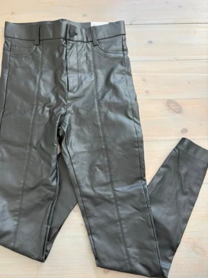 Leggins Zara nytt  - Snygga svarta leggings i fuskskinn med markerade sömmar framtill. Hög midja och bälteshällor för extra stilpoäng. Perfekta för dig som vill ha en edgy look och gillar trendiga detaljer.